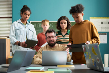 Caucasian man teaching diverse group of teenagers using digital tablets and laptops in classroom, students standing around teacher engaging with technology and learning