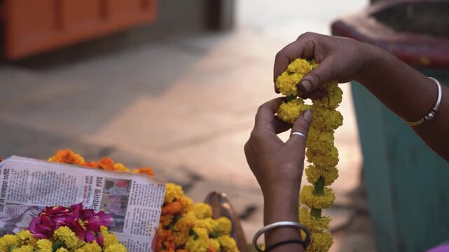 Close-up of a woman's hands stringing fresh yellow marigold flowers (genda phool) into a traditional garland for a Hindu festival like Diwali | Streen vendor