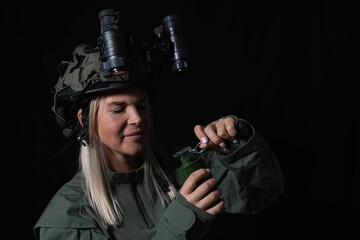 A female soldier in a tactical helmet with night vision goggles prepares a hand grenade against a black background.