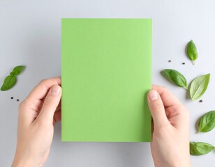 Hands holding a blank lime green sheet of paper surrounded by fresh basil leaves and peppercorns on a light gray surface