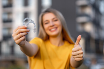 Smiling woman holding clear dental aligners and showing thumbs up outdoors