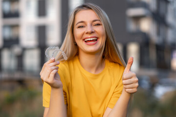 Smiling woman holding clear aligners and showing thumbs up outdoors
