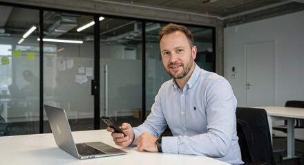 Fototapeta premium Businessman using smartphone at a desk with laptop in a modern open-office environment