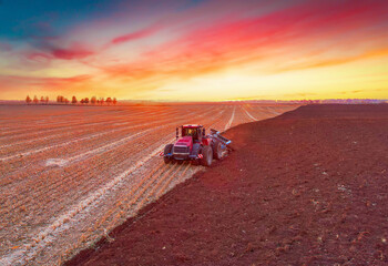 Fototapeta premium Plowing field on sunset. Red Tractor with Plough on Plowed. Ploughing, Soil Tillage. Agricultural Tractor on Cultivation Field for Sowing Seeds. Grain deal, Tractor on Field Cultivating in Spring.