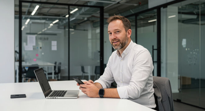 Businessman using smartphone at a laptop on a clean desk in a modern office - Powered by Adobe