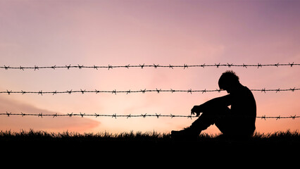 Solitary Silhouette: A lone figure sits dejectedly beside a barbed wire fence, silhouetted against...
