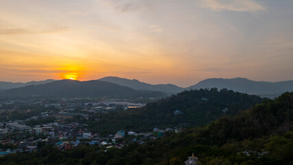 A scenic view of a town nestled among lush green mountains at sunset, with a temple structure perched on the hillside and a vibrant orange sky creating a warm, tranquil atmosphere.