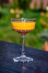Bright orange cocktail in vintage crystal glass against summer garden background on wooden table, closeup. Delicious orange alcoholic drink for relaxation on a sunny warm day