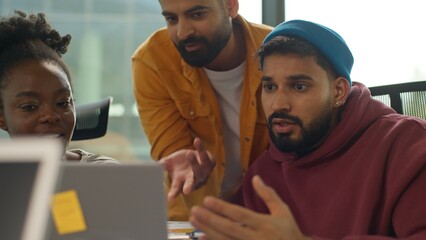 Three diversity colleagues workers in casual office arguing about document data looking in laptop multiracial informal small business team emotional talking discuss work marketing results smiling