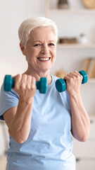 Home training concept. Strong senior woman doing exercises with dumbbells indoors. Cheerful mature lady working out her arm muscles, keeping fit, leading healthy lifestyle during covid-19 isolation