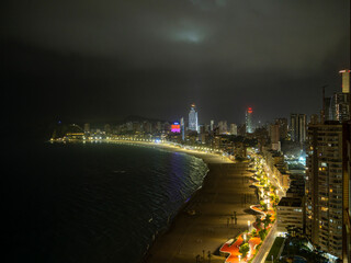 Night view of Benidorm, Spain, with illuminated skyscrapers, a curving coastline, and a glowing beachfront promenade. The vibrant lights contrast beautifully against the dark sea and sky.
