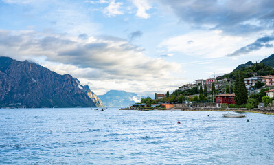 Dramatic mountains surround Lake Garda under a cloudy sky, with sunlight breaking through to highlight the rocky landscape. The calm water contrasts with the powerful peaks of northern Italy.