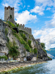 A medieval castle in Malcesine stands atop rugged cliffs overlooking Lake Garda. Its stone tower and crenellated walls contrast beautifully with the sky and shimmering blue water below.
