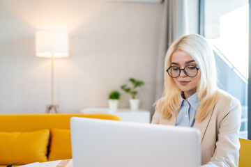 Blonde businesswoman in eyeglasses using laptop in cozy modern office interior for remote work, freelancing, home office, digital communication, career success, online education, productivity.