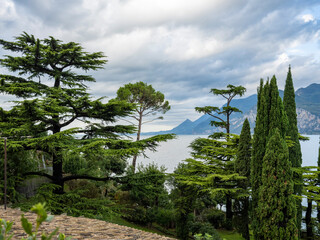 Ancient castle walls rise above Lake Garda, surrounded by tall cypress trees and lush greenery. The view opens to dramatic cliffs and blue water under a cloudy sky in northern Italy.