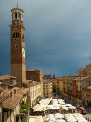 Torre dei Lamberti rises over Verona&rsquo;s lively Piazza delle Erbe, where people gather under white market umbrellas, framed by colorful historic buildings and a dramatic stormy sky.