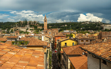 A panoramic view of Verona&rsquo;s historic center with terracotta rooftops, the Church of St. Anastasia, and Castel San Pietro in the distance under a dramatic sky.