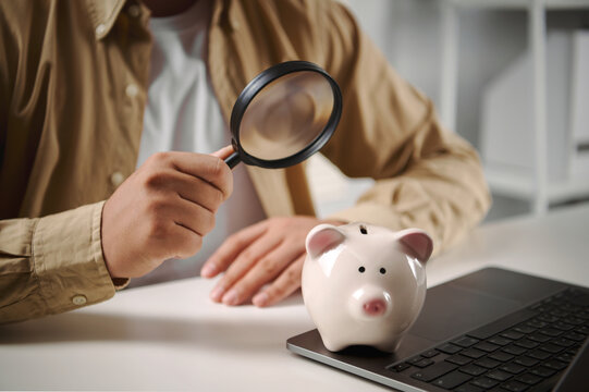 Man examining piggy bank with magnifying glass on laptop concept of financial audit money saving analysis budget planning and economic investment evaluation