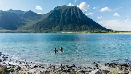 Two individuals wade into the clear blue waters of Lofoten, Norway. Surrounded by majestic mountains and vibrant greenery, this serene summer day invites adventure and relaxation in nature embrace. © Fokke Baarssen