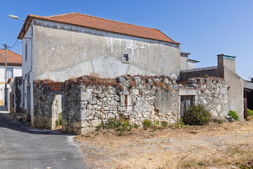 A side view of a derelict Portuguese building with crumbling stone walls, a red-tiled roof, and dry grass in front, showcasing its state of neglect