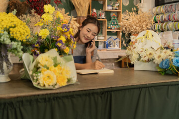 Asian female florist in apron smiling while designing a beautiful floral arrangement with her colleague in a flower shop. Concept of teamwork, creativity, floral business, and joyful workplace.