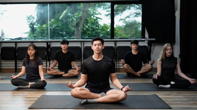 Young fit multiracial people in sportswear sitting in row on floor mat in lotus padmasana position, meditating with closed eyes and folded in mudra gesture fingers on knees at yoga group training.