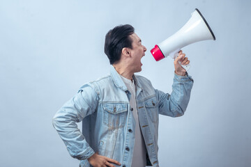 Asian man in denim jacket shouting through a white megaphone with an expressive face, symbolizing...