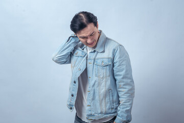 Fototapeta premium Asian man in denim jacket holding shoulder with painful face, standing in studio against plain light background, expressing shoulder ache, injury, discomfort, and health problem