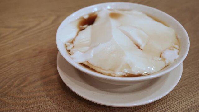 A bowl of traditional Chinese dessert tofu pudding, also known as tau foo fah or douhua, served with sweet syrup. Silky smooth soy bean curd dessert in a white bowl on wooden table.