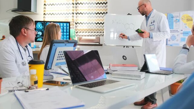 Bald pharmaceutical representative in white coat writes with marker on whiteboard holding phone during hospital seminar. Medical staff listen to lecture about new drug and healthcare innovations.