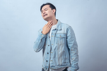 Asian man in denim jacket clutching his throat with distressed expression, standing in studio against plain light background, showing choking, breathing difficulty, and pain