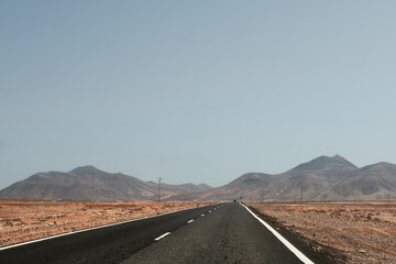 Straight road cutting through the desert of Fuerteventura, leading towards arid mountains under a clear sky.
