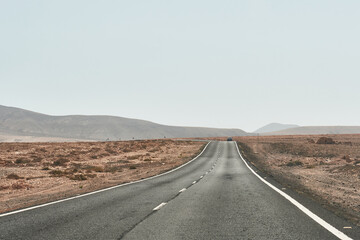 Photograph of a solitary asphalt road across the dry desert landscape of Fuerteventura, with low mountains in the distance.