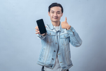 Fototapeta premium Asian man in denim jacket holding smartphone with blank screen and giving thumbs up, showing excited expression on plain studio background