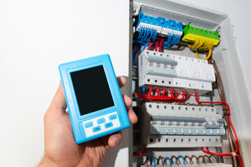 Hand holding portable measuring device with blank screen near open electrical panel, ready for design mockup.
