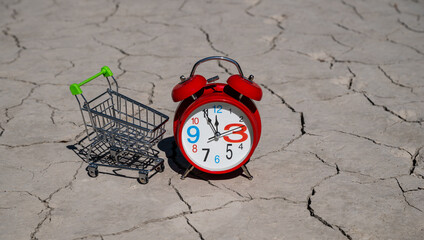 An alarm clock and a miniature supermarket shopping cart on hot, cracked clay