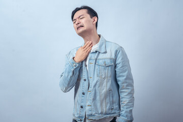 Fototapeta premium Asian man in denim jacket clutching his throat with distressed expression, standing in studio against plain light background, showing choking, breathing difficulty, and pain