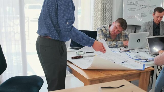 Frustrated bald manager in glasses scolding architects and office workers during stressful project meeting. Team with laptops, blueprints and plans shows workplace conflict and pressure.