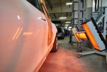 A white car with side damage stands in a repair shop, illuminated by red infrared heat lamps for paint curing, surrounded by tools and other vehicles in a busy garage setting.