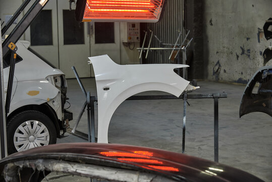 A white car fender under a red heat lamp in a repair shop, with tools, paint splatter, and a damaged vehicle nearby, showing an active auto bodywork and restoration process. - Powered by Adobe