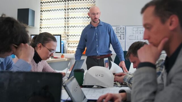 Bald manager in glasses emotionally arguing with architects during tense meeting in modern office. Team working with laptops, blueprints and project plans, showing conflict and workplace stress.
