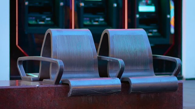Chairs placed in front of brightly lit ATMs during evening time. Loungers standing still near cash dispensers on quiet urban street. Seats appearing vacant by glowing machines in city twilight