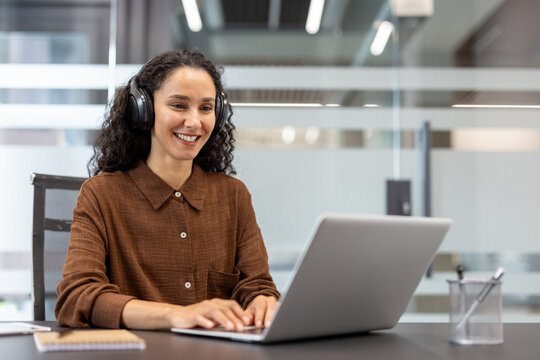 Woman wearing headphones smiling while typing on a laptop, connecting remotely for a business meeting or online work in a modern corporate office environment - Powered by Adobe