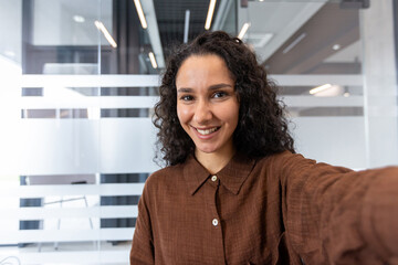 Young professional woman with curly hair smiling broadly, holding out her arm to take a selfie in a...