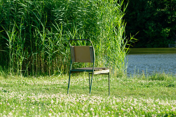 An old chair sits peacefully on the green grass near a lake on a sunny day.