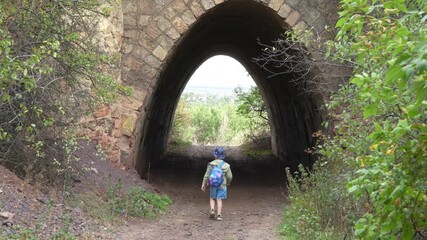 4K video of a boy with a backpack walking into a stone tunnel on a forest path. Concept of childhood adventure, exploration and journey - Powered by Adobe