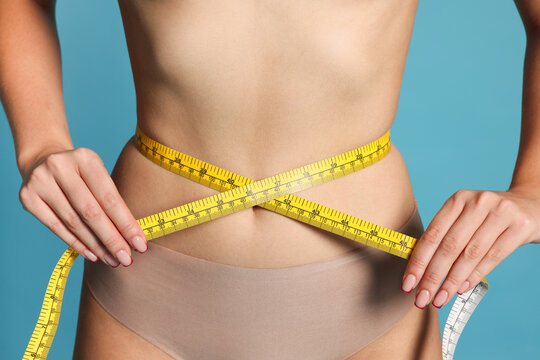Young woman measuring her flat belly with tape on light blue background, closeup