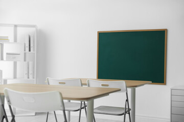 Blank green chalkboard, desks, chairs and shelving unit in classroom