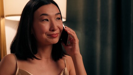 Close up positive peaceful Asian woman Korean female Chinese lady Japanese elegant style at luxury home room evening indoors talking mobile phone smiling talk call smartphone communicate conversation