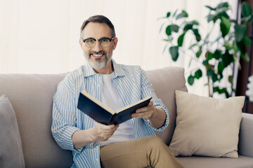 Smiling mature man reading a book on a couch at home near bright window, enjoying a relaxed and productive moment